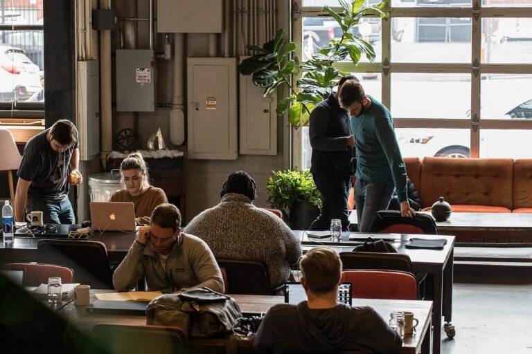 Several people working on laptops and documents at long wooden tables in a shared coworking space with large industrial windows and indoor plants