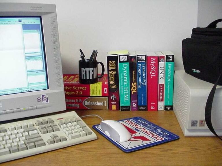 A desktop computer workstation with a CRT monitor, keyboard, mouse on a mousepad, a mug holding pens, and a row of technical programming and web development books including titles on SQL Server, Dynamic HTML, MySQL, UNIX, and JavaScript, on a wooden desk