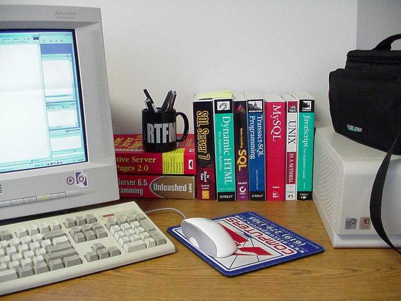 A desktop computer workstation with a CRT monitor, keyboard, mouse on a mousepad, a mug holding pens, and a row of technical programming and web development books including titles on SQL Server, Dynamic HTML, MySQL, UNIX, and JavaScript, on a wooden desk
